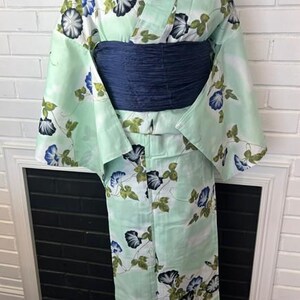 Op de afbeelding: Een licht muntgroene yukata met een herhalend patroon van blauwe en zwarte ochtendgloriebloemen met groene bladeren. Het kledingstuk heeft wijde mouwen en is in de taille vastgezet met een brede, getextureerde, donker marineblauwe obi-ceintuur.