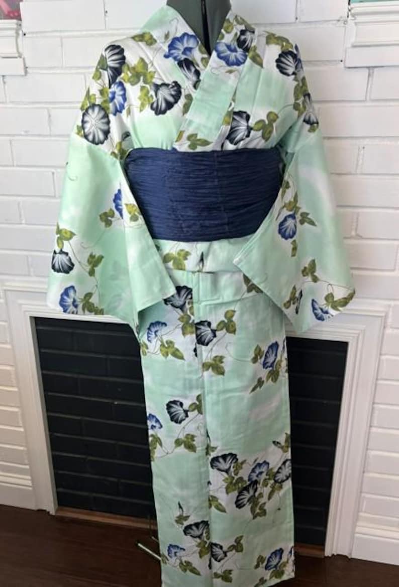 Op de afbeelding: Een licht muntgroene yukata met een herhalend patroon van blauwe en zwarte ochtendgloriebloemen met groene bladeren. Het kledingstuk heeft wijde mouwen en is in de taille vastgezet met een brede, getextureerde, donker marineblauwe obi-ceintuur.
