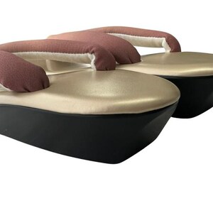 May include: A pair of traditional Japanese geta sandals. The sandals feature a black base, a gold-colored top, and a brown fabric thong. The thong is padded with white fabric. The sandals are elevated.