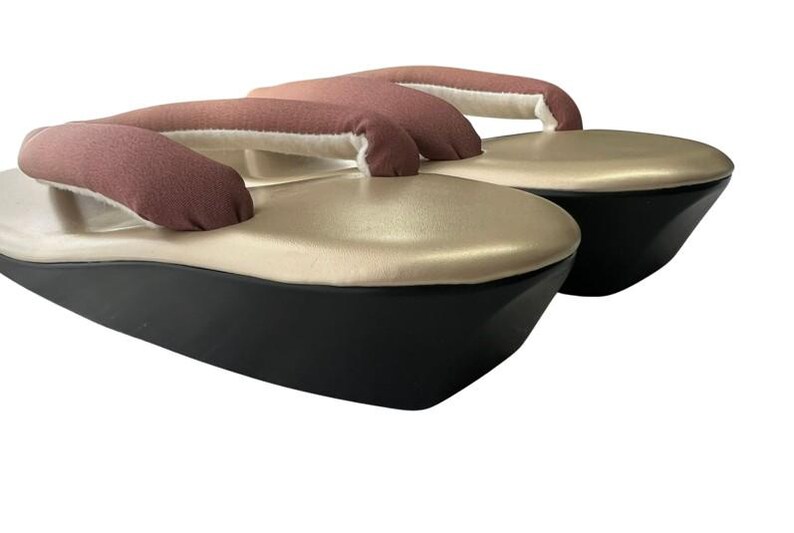 May include: A pair of traditional Japanese geta sandals. The sandals feature a black base, a gold-colored top, and a brown fabric thong. The thong is padded with white fabric. The sandals are elevated.