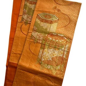 Op de afbeelding: Een oranje textiel met een herhalend ruitpatroon en sierlijke, geometrische ontwerpen in groen, rood en goud. Het textiel heeft gestileerde doosachtige motieven en decoratieve kwasten.