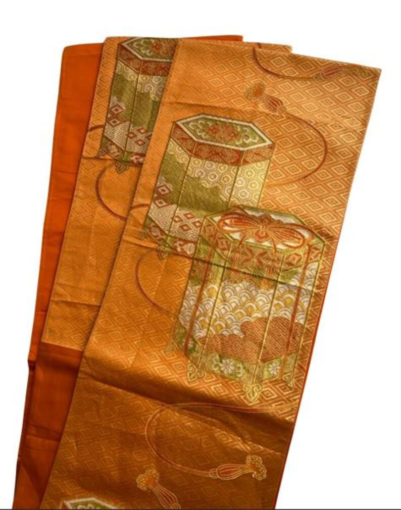 Op de afbeelding: Een oranje textiel met een herhalend ruitpatroon en sierlijke, geometrische ontwerpen in groen, rood en goud. Het textiel heeft gestileerde doosachtige motieven en decoratieve kwasten.