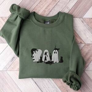 Peut inclure: Un sweat-shirt vert avec un motif brodé de quatre fantômes et de chats noirs. Les fantômes sont blancs et ont un design simple. Les chats sont noirs et sont dans une pose ludique.