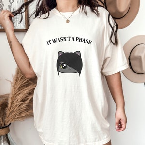 May include: White oversized t-shirt with the text "It wasn't a phase" and a cartoon black cat with a black emo hairstyle.