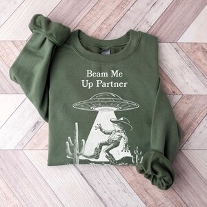 以下が含まれることがあります： ビーム・ミー・アップ・パートナーという文字と、UFO、サボテン、カウボーイの宇宙人の白いグラフィックが描かれた緑色のスウェットシャツです。