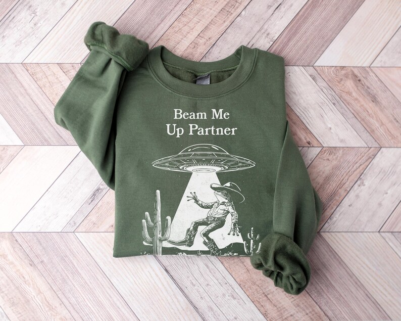 以下が含まれることがあります： ビーム・ミー・アップ・パートナーという文字と、UFO、サボテン、カウボーイの宇宙人の白いグラフィックが描かれた緑色のスウェットシャツです。