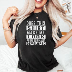 Peut inclure: Un t-shirt noir à manches courtes avec un texte blanc qui dit : DOES THIS SHIRT MAKE ME LOOK PROFESSIONALLY DEVELOPED. Le lettrage est disposé dans un format de bloc empilé avec un aspect texturé et usé sur le devant au centre.