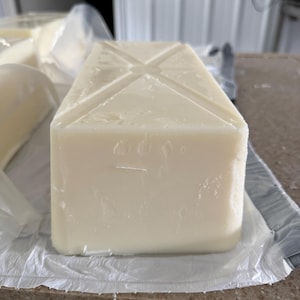 May include: A large, rectangular block of white soap with a cross-shaped design on the top. The soap is sitting on a white, crinkled paper. Another wrapped bar of soap is visible in the background.