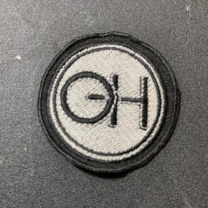May include: Round embroidered patch with a gray center and black border. The patch features the letters "OH" in black, with the "O" overlapping the "H". The patch is likely for clothing or accessories.