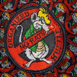 May include: Embroidered patch with a red border and the words "ORGANIZED CRUSTLE" and "NO RATS" in a circle. The patch features a cartoon rat with a yellow hat and green clothing, crossed out with a red line. A pile of similar patches is in the background.