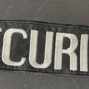Peut inclure: Un écusson rectangulaire noir avec le mot "SECURITY" brodé en lettres majuscules blanches. L'écusson a une bordure noire et est conçu pour être cousu sur des vêtements ou un uniforme.