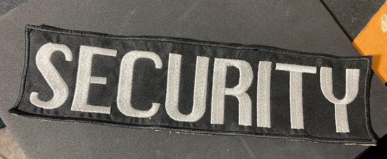 May include: A black rectangular patch with the word "SECURITY" embroidered in white capital letters. The patch has a black border and is likely designed to be sewn onto clothing or a uniform.
