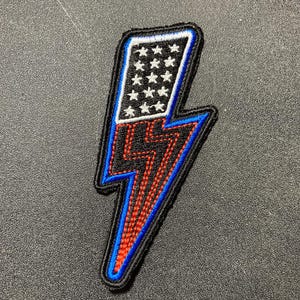 May include: Embroidered patch in the shape of a lightning bolt. The top section features a white field with white stars on a blue border. The bottom section is red and black with a blue border. The patch has a black border.