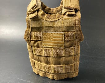 Puede incluir: Un chaleco táctico de color canela con un parche de la bandera estadounidense. El chaleco presenta múltiples correas y un patrón de rejilla para sujetar equipo adicional. El chaleco está hecho de una tela duradera y texturizada.