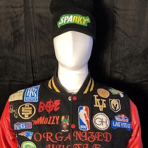 May include: A black varsity jacket with red sleeves and a black beanie with the word "Sparky" embroidered in green and yellow. The jacket is covered in patches, including a basketball logo, a "Mozzy" patch, a "Royce" patch, and a "Las Vegas" patch. The jacket also has the words "Organized Hustle" embroidered on the front.