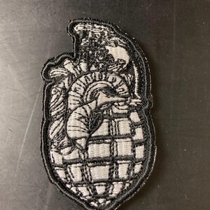 May include: A gray and black embroidered patch featuring a stylized heart design within a grenade shape. The patch has a black border and intricate detailing, suitable for jackets, bags, or other fabric items.