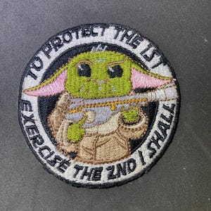 Puede incluir: Parche bordado circular con un personaje verde alienígena sosteniendo un bláster. El parche tiene un diseño circular con el texto "TO PROTECT THE 1ST EXERCISE THE 2ND I SHALL" en letras blancas sobre fondo negro.