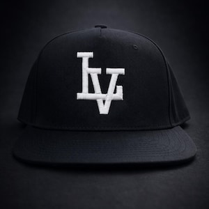 Peut inclure: Casquette de baseball noire avec une visière plate. Le chapeau présente les lettres blanches brodées "LV" sur le devant. Le chapeau est posé sur un fond sombre.