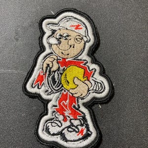 Könnte beinhalten: Gestickter Aufnäher mit einer Cartoonfigur, die einen weißen Hut mit rotem Blitz trägt. Die Figur hält einen gelben Ball und hat rote Akzente auf der Kleidung. Der Aufnäher hat einen schwarzen Rand.