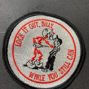 Könnte beinhalten: Runder Aufnäher mit rotem gesticktem Text "LOCK IT OUT, BILLY WHILE YOU STILL CAN". Das Design zeigt eine Cartoonfigur in Rot, die eine andere in Schwarz angreift. Der Aufnäher hat einen grauen Hintergrund und einen schwarzen Rand.