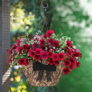 May include: A hanging basket overflowing with vibrant red petunias and delicate white flowers. The basket is made of a burlap-like material with a black damask pattern and a black bow. The basket is suspended by a metal hanger.