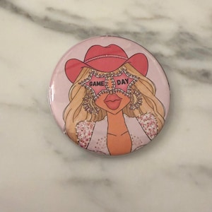 May include: A circular pinback button featuring a graphic illustration of a blonde person wearing a pink cowboy hat and star-shaped sunglasses with the text GAME DAY printed on the lenses. The person has long blonde hair and wears a white top.