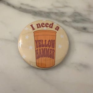 May include: A circular pin-back button with a light beige background featuring white stars. The center displays an orange plastic cup graphic with the text I need a YELLOW HAMMER printed in dark red, bold, retro-style lettering across the cup.