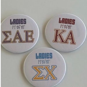 Puede incluir: Tres botones redondos blancos con texto. Cada botón tiene las palabras "LADIES PREFER" encima de letras griegas. El primer botón tiene "ΣAE" en granate, el segundo tiene "KA" en granate y el tercero tiene "ΣX" en azul y dorado.