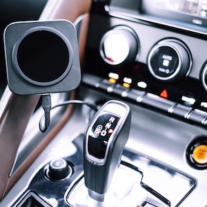 May include: Interior view of a car's dashboard, featuring a black wireless charging pad with a circular charging area. The dashboard includes climate control knobs, a gear shift, and a start button. The car's interior has brown leather accents.