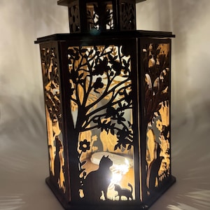 May include: A wooden lantern with a hexagonal shape and intricate cutouts of trees, flowers, and cats. The lantern is lit from within, casting a warm glow.