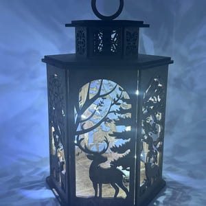 May include: A wooden lantern with a hexagonal shape and a deer silhouette inside. The lantern has a decorative top with a ring and intricate cutouts. The deer is surrounded by trees and other woodland elements. The lantern is lit from within, creating a warm glow.