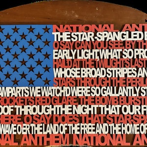 Puede incluir: Un letrero de madera con forma de bandera con las palabras "National Anthem" y la letra de "The Star-Spangled Banner" impresas en letras rojas, blancas y azules. El letrero tiene un fondo azul con estrellas blancas.