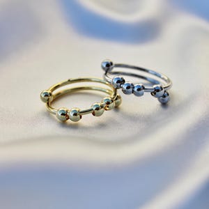 Può includere: Due anelli, uno color oro e uno color argento, ciascuno caratterizzato da una sottile fascia con cinque piccole perline rotonde e lisce posizionate per scorrere lungo il filo metallico. Gli anelli hanno una finitura lucida e riflettente.