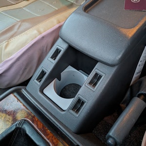 May include: A grey car centre console with a cup holder, four rectangular slots, and a grey handbrake lever. The console is made of plastic and has a textured surface.