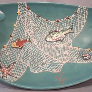 May include: A teal ceramic bowl with a nautical theme. The bowl features a white fishing net design with painted fish, a starfish, and a seashell. The rim has curved handles. The bowl is approximately 30 cm in diameter.