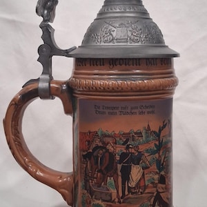 May include: A brown ceramic beer stein with a pewter lid and handle. The lid is topped with a sculpture of a rider on a horse. The body of the stein features a painted scene of people and buildings, with German text.