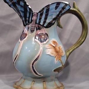 May include: A ceramic pitcher with a butterfly design. The butterfly wings are blue and black, and the body is purple. The pitcher is light blue with a flower and leaf design. The handle is green.
