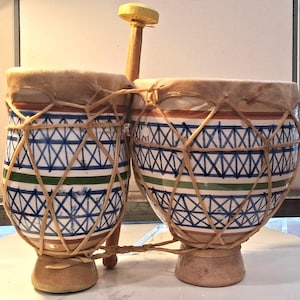 May include: A pair of ceramic bongo drums connected by a wooden rod, featuring a white base with hand-painted blue cross-hatch patterns and horizontal green and brown stripes. The drum heads are made of light tan animal skin secured with twine.