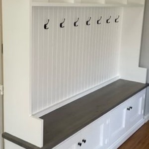 Può includere: Panca d'ingresso in legno bianco con piano grigio, mensole aperte e ganci. La panca ha due ante bianche con pomelli neri.