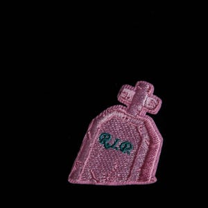 Op de afbeelding: Een roze geborduurde patch in de vorm van een grafsteen met een kruis erop. De patch heeft de letters "R.I.P." geborduurd in groen. De achtergrond is zwart.