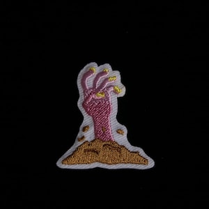 May include: Embroidered patch showing a pink zombie hand rising from the ground. The hand has yellow fingernails and is stitched on a white background with brown soil. The patch is set against a black background.
