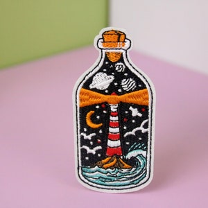 May include: Embroidered patch of a lighthouse in a bottle with a cork top. The bottle is filled with a black background with white stars, a crescent moon, and a few white birds. The lighthouse is red and white striped with a yellow beam of light. The bottom of the bottle shows blue waves.