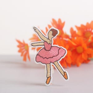 May include: Embroidered patch of a ballerina in a pink tutu, arms raised. The patch has a white border and is set against a backdrop of orange flowers. The ballerina's hair is brown, and she wears orange ballet slippers.