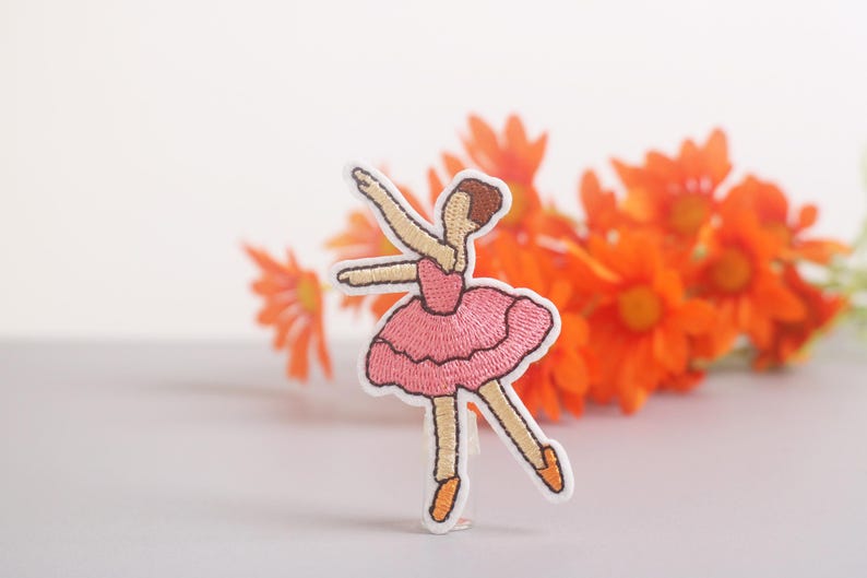 May include: Embroidered patch of a ballerina in a pink tutu, arms raised. The patch has a white border and is set against a backdrop of orange flowers. The ballerina's hair is brown, and she wears orange ballet slippers.