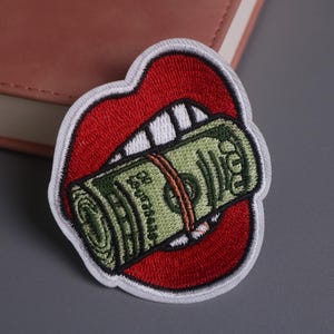 Puede incluir: Parche bordado que representa una boca roja con dientes blancos, sosteniendo un rollo de dinero verde. El parche tiene un borde blanco. El rollo de dinero está asegurado con una banda marrón. El parche está sobre una superficie gris.