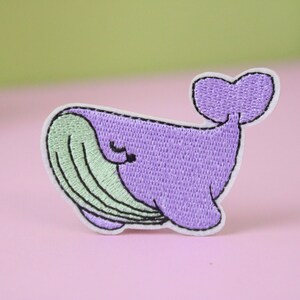 May include: Purple and green embroidered whale patch. The whale has a smiling face and a closed eye.