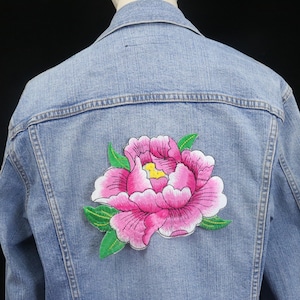 Peut inclure: Une veste en jean bleu clair avec un patch brodé de fleurs roses et vertes au dos.