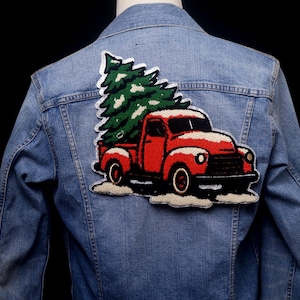 May include: A denim jacket with a large embroidered patch featuring a red vintage truck carrying a green Christmas tree. The truck is covered in snow, creating a festive holiday design.
