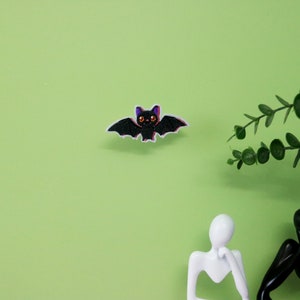 Puede incluir: Aplique en forma de murciélago negro con ojos naranjas y detalles morados, sobre un fondo verde claro. Decoración temática de Halloween. Una ramita de hojas verdes y dos figuras humanas abstractas también están en la imagen.