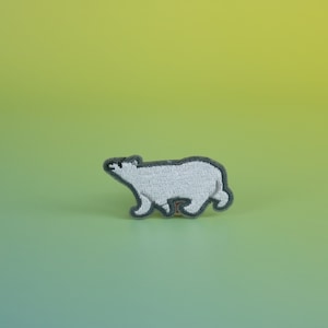 以下が含まれることがあります： 白いくまのデザインの刺繍パッチ。くまは白い色で、灰色の輪郭があり、右方向に歩いています。パッチは緑と黄色のグラデーションの背景にあります。